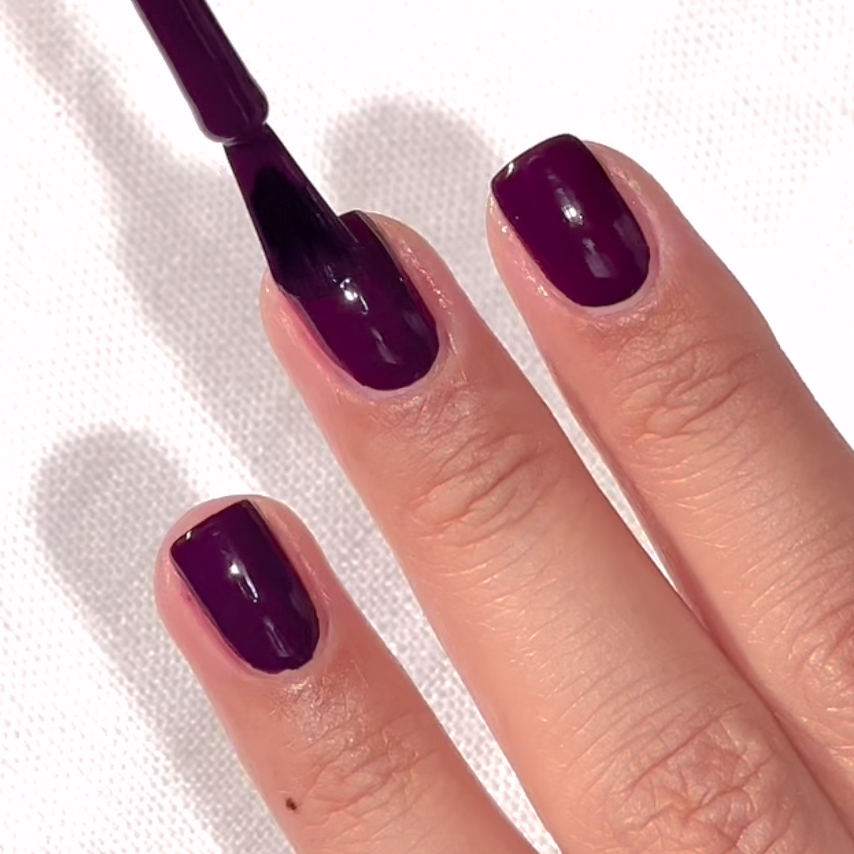 A close-up of a hand with short, neatly trimmed nails being painted with glossy dark purple nail polish on a white textured surface.