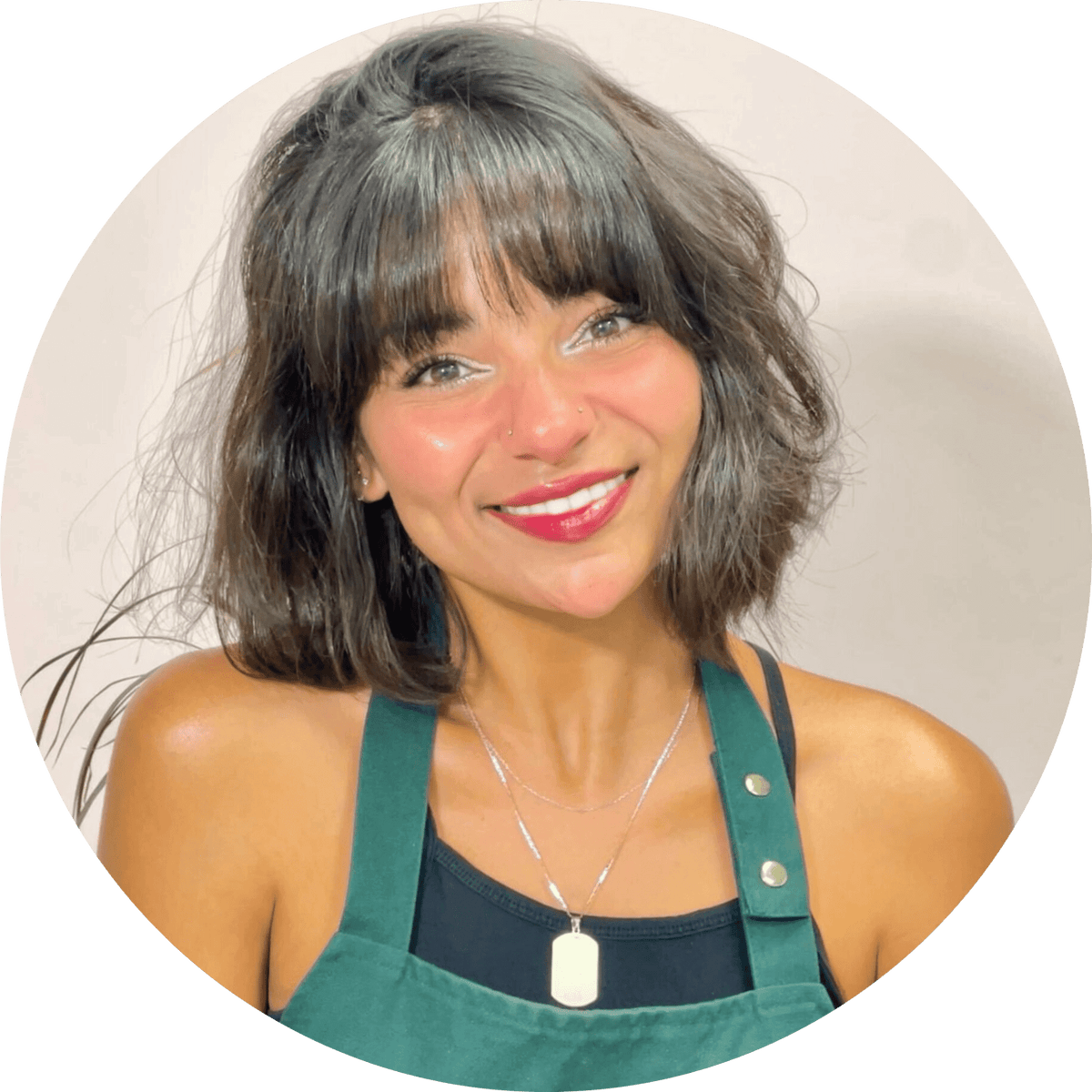 A woman with short, wavy dark hair and bangs smiles at the camera. She is wearing a green apron over a black top and a silver necklace with a rectangular pendant. The background is light and plain.
