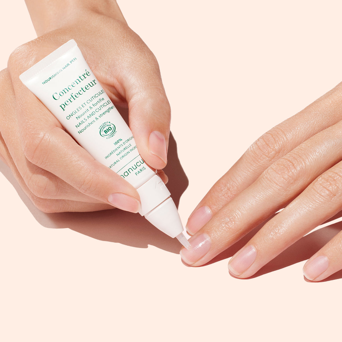 A hand applies Manucurist’s Stronger Nails Duo onto another’s nails against a pale pink background, repairing and perfecting nails for a healthy look.