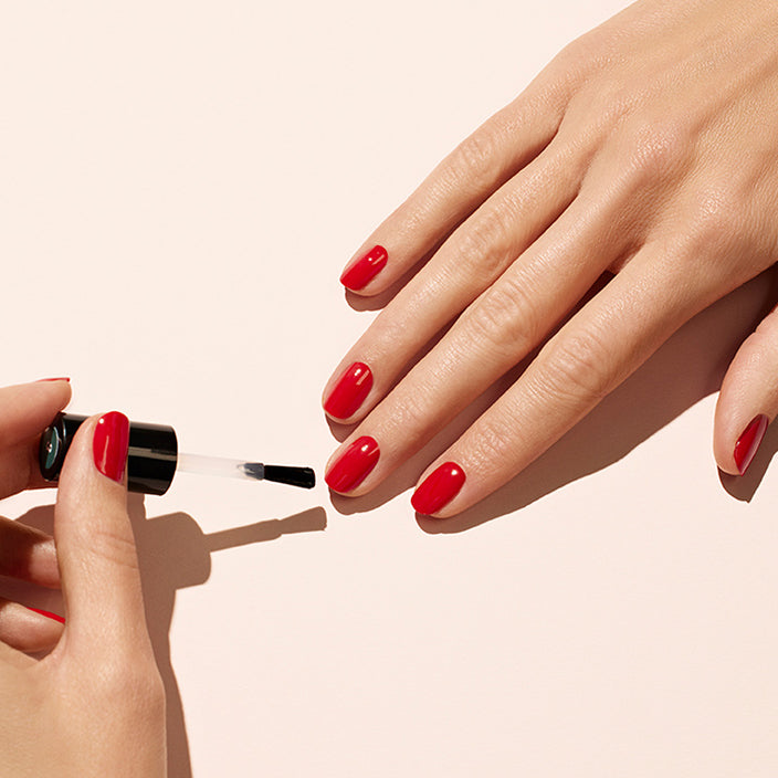 A close-up of one hand applying red nail polish to the nails of the other hand against a light beige background. The nails are neatly shaped and all fingernails are being polished.