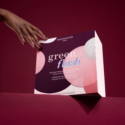 A hand with painted nails holds the Manucurist Starter Kit Gift Set (TEST COFFRET) featuring pink, white, and purple circles against a dark pink background. The semi-permanent manicure kit offers acetone-free removal.