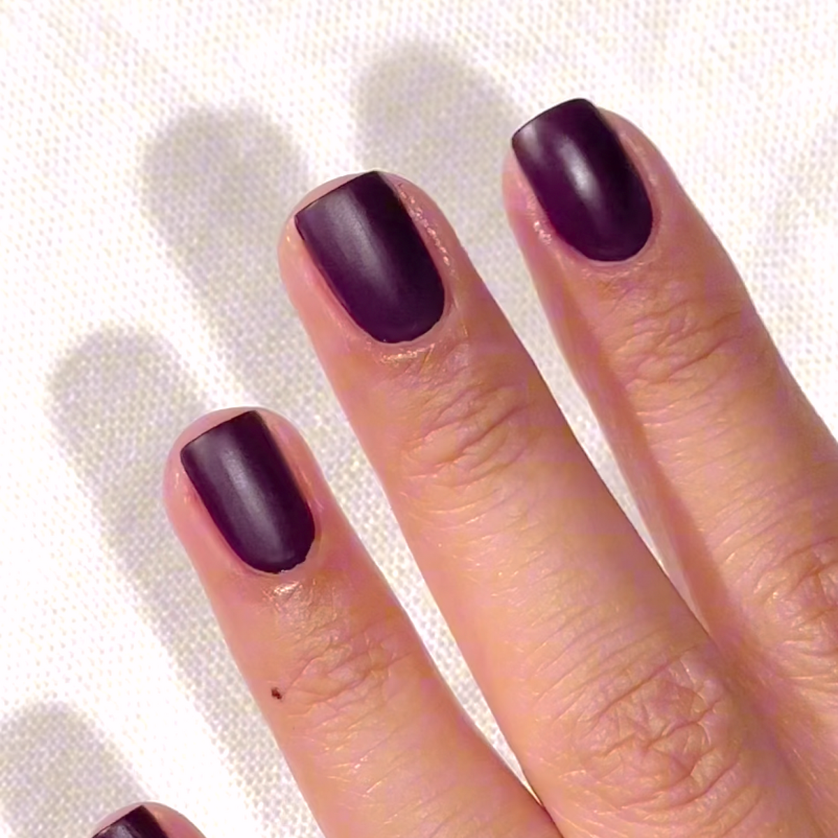 A hand with neatly manicured nails painted in a matte dark purple shade, resting on a white textured fabric background.