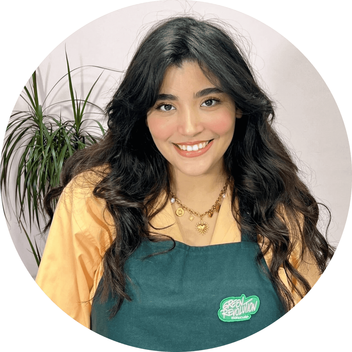 A woman with long dark hair wearing a green apron and a yellow shirt smiles at the camera. She stands in front of a light background with a leafy green plant to her side.