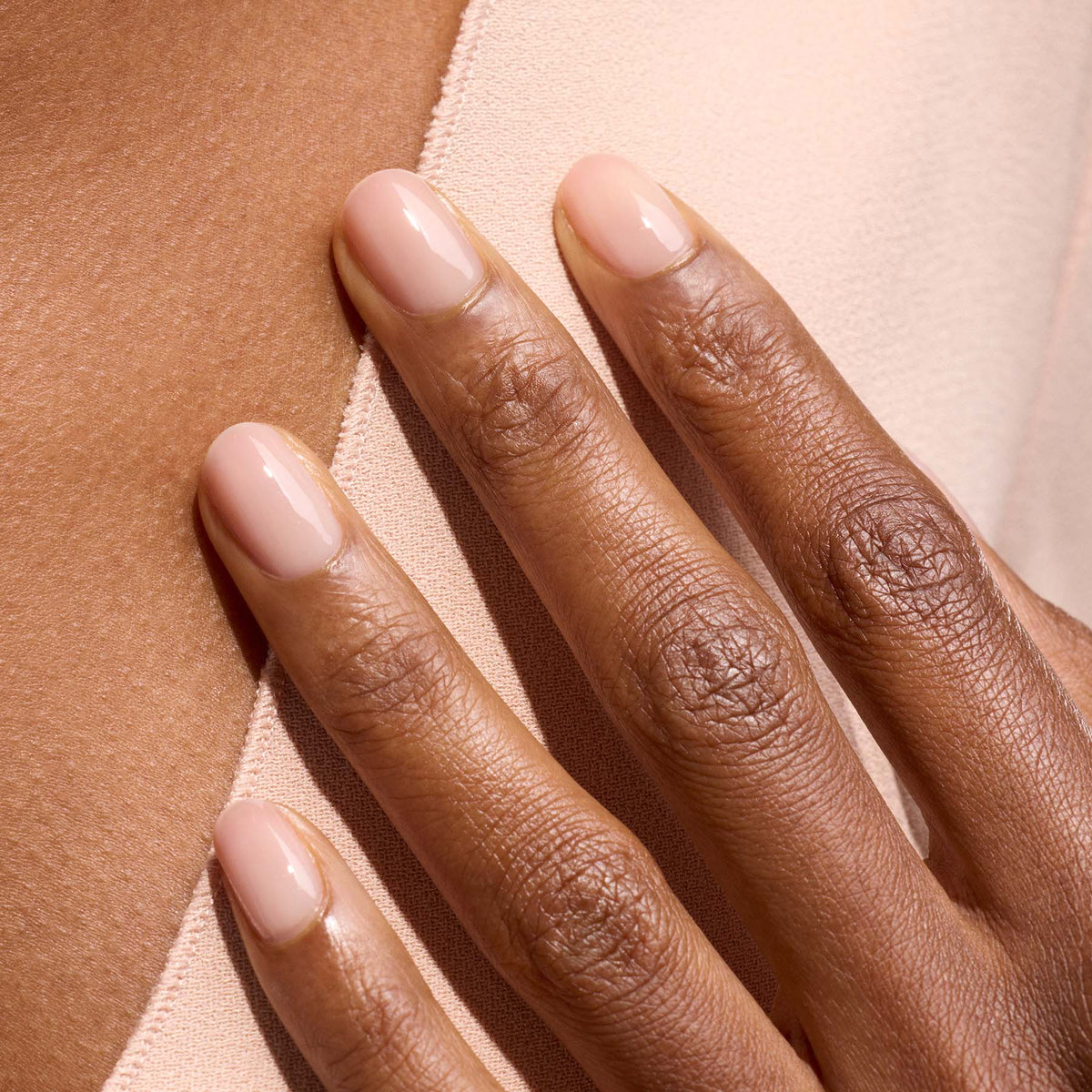 Gros plan sur une main aux ongles courts et ovales vernis avec Pale Rose de Manucurist, un vernis rose pastel translucide, posée sur un tissu beige clair et dévoilant une manucure élégante et naturelle.