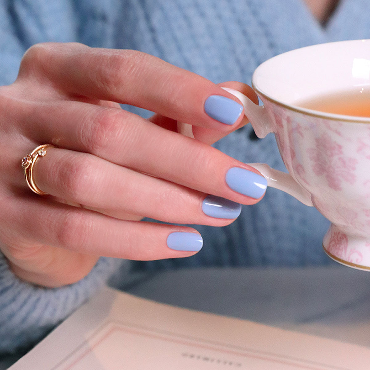Une main aux ongles soigneusement manucurés et vernis avec le coffret You  Me Under the Sea Green Flash™ de Manucurist tient une tasse de thé blanche à motif floral rose. La personne porte une bague dorée, un pull bleu, et un livre ouvert est partiellement visible.