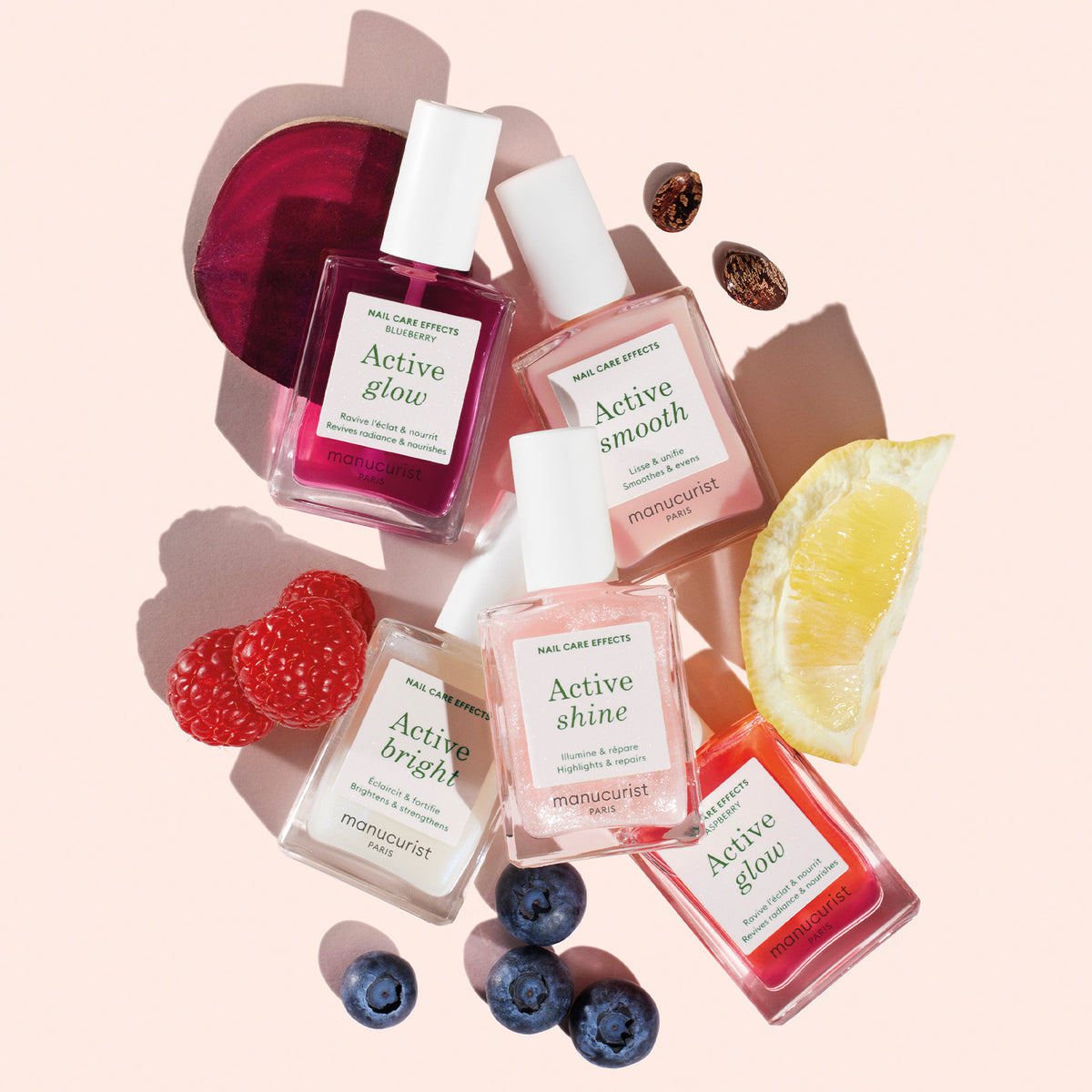 Five bottles from the Manucurist Super Ultimate Active Manicure Kit are displayed on a light pink surface, surrounded by raspberries, blueberries, sliced lemon, cranberries, and a slice of beetroot.