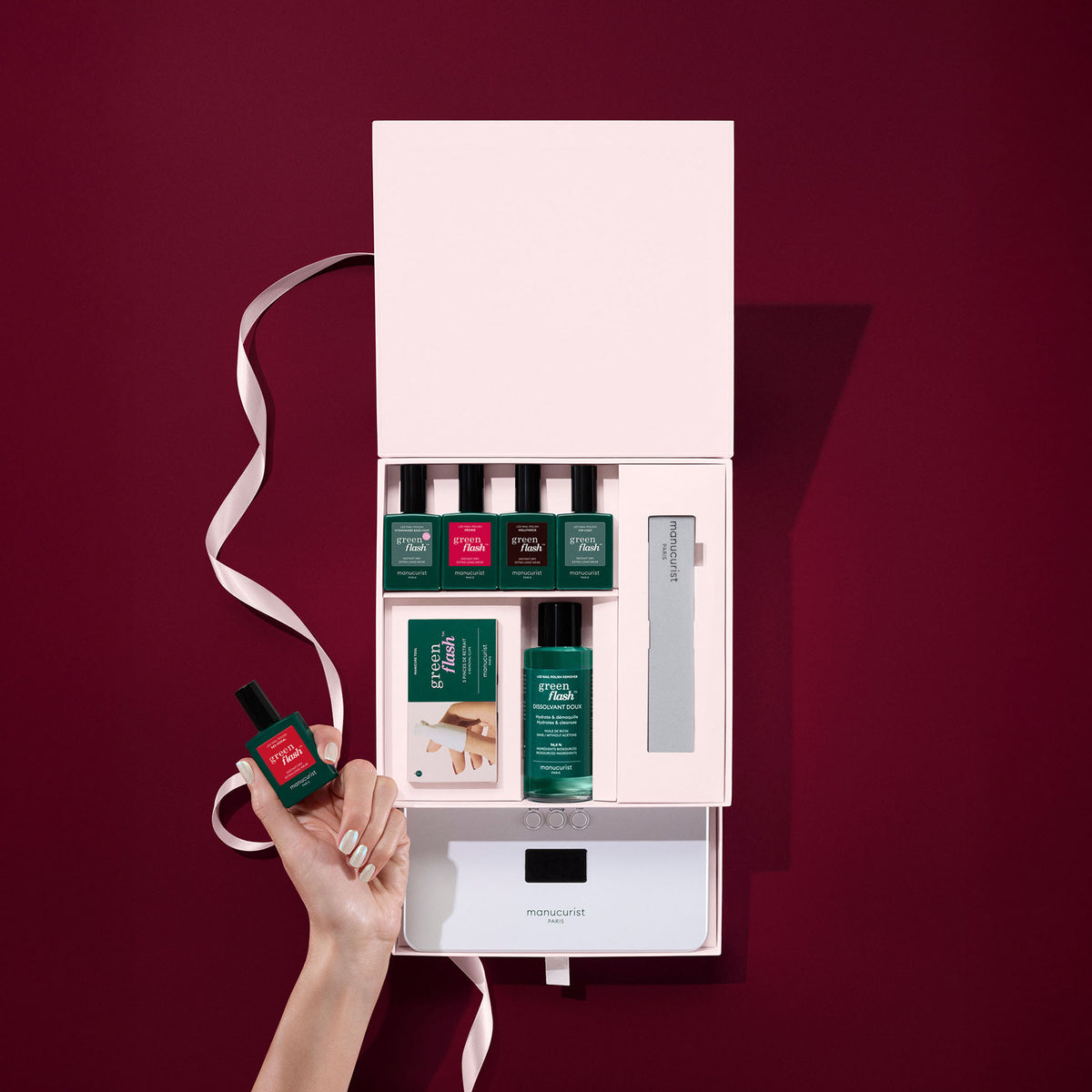 A hand holds a red polish bottle over the Manucurist Starter Kit Gift Set (TEST COFFRET), featuring four Green Flash semi-permanent bottles, a nail file, and tools, set against a burgundy background with a white ribbon.