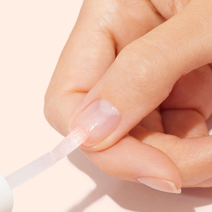 A close-up of a hand applying Manucurist Strong Glow Trio to a fingernail with a brush against a light beige background, emphasizing gentle nail care and treatment for damaged nails.