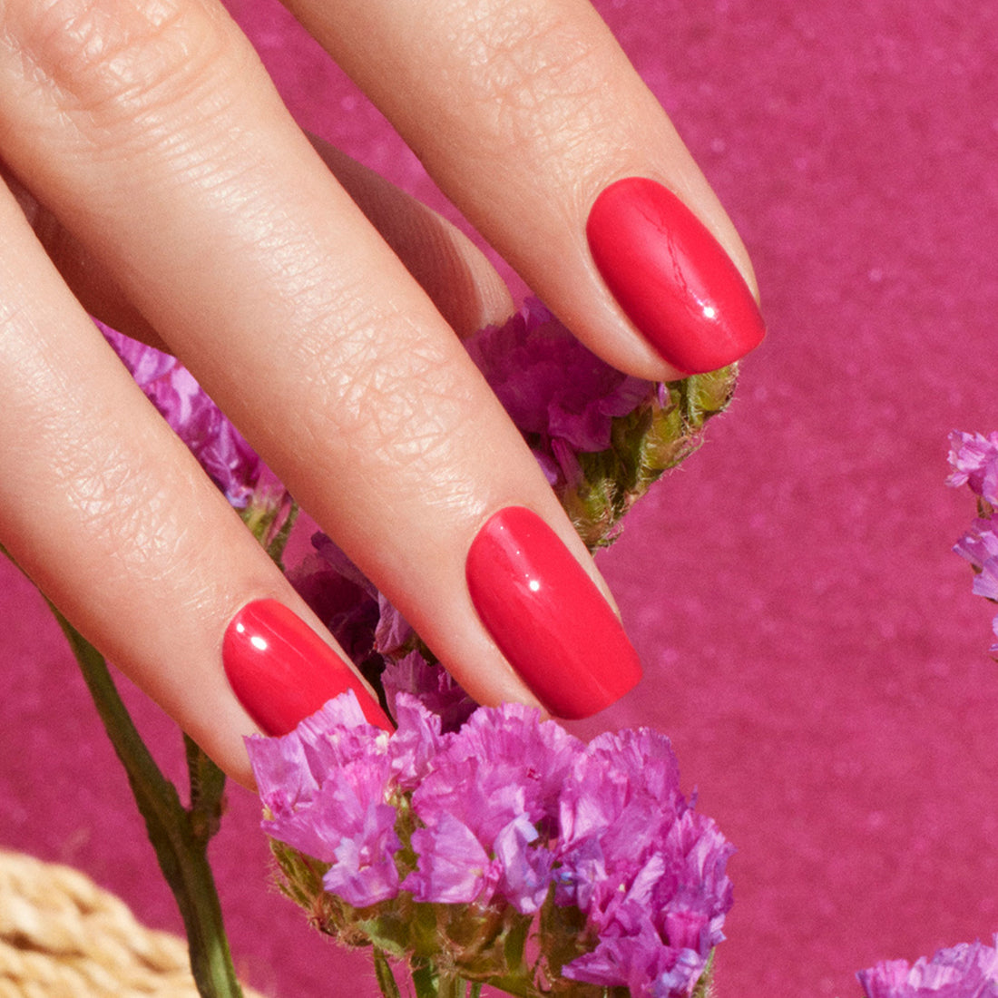 Gros plan sur une main portant la teinte Bougainvillea de Manucurist, un vernis à ongles rose rougeâtre brillant, tenant de petites fleurs de bougainvillier sur un fond rose éclatant.