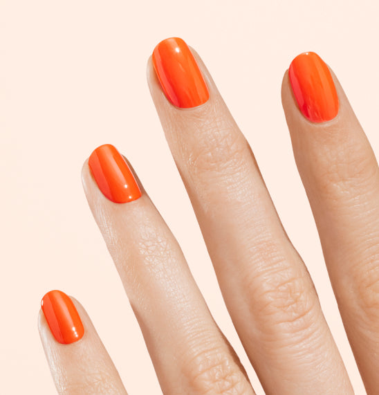 A close-up of a hand with four fingers visible, each with neatly manicured nails painted in shiny bright orange nail polish against a light beige background.
