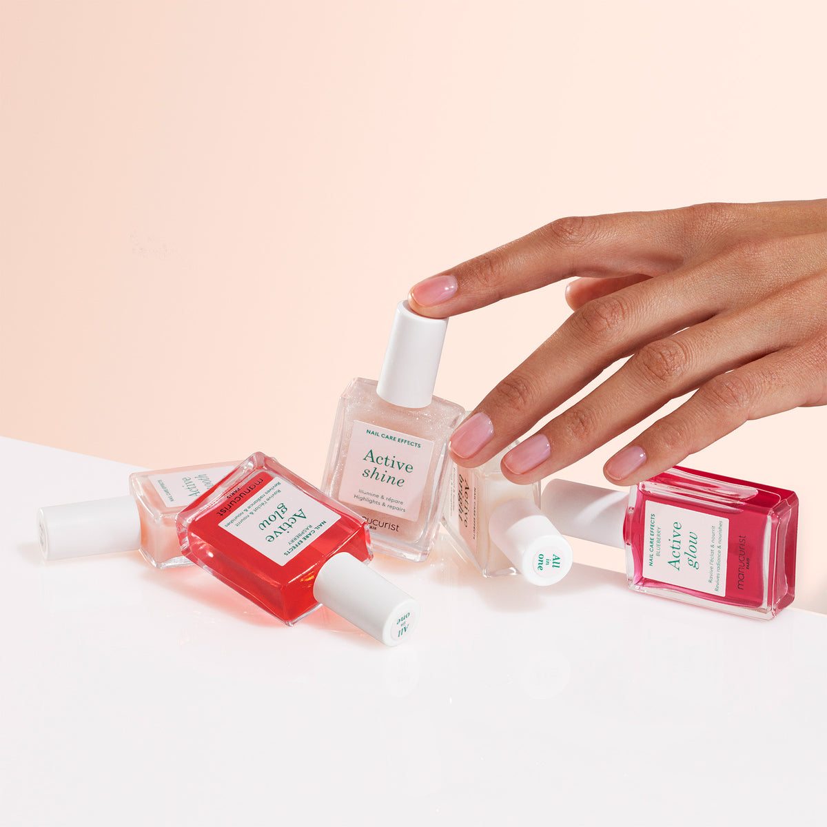 A hand with manicured nails reaches for a clear nail polish bottle labeled Active Shine among several bottles of red and pink nail polish on a white surface with a soft peach background.