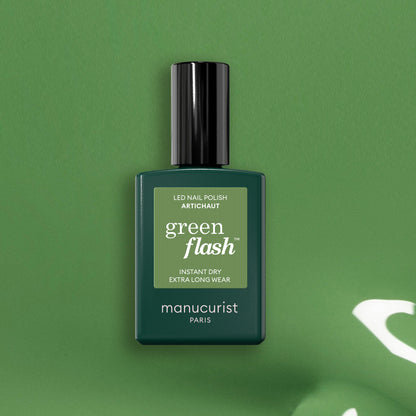 Un flacon de vernis LED Manucurist Artichoke Green Flash repose sur un fond vert assorti. Le flacon rectangulaire arbore un bouchon noir et un texte blanc, évoquant la fraîcheur luxuriante d&