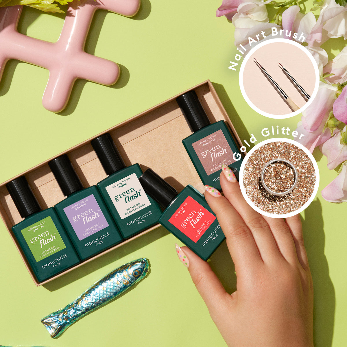 A hand selects a bottle of Manucurist Glitter in Love nail polish from a set of five in a box, with a toy fish nearby. Inset circles display nail art brushes and biodegradable glitter on a neutral background.