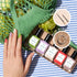 A hand with multicolored nails rests on a striped cloth next to five green-label Manucurist "Glitter in Love" nail polish bottles, biodegradable glitter, thin nail art brushes, and a green mesh bag of lettuce.