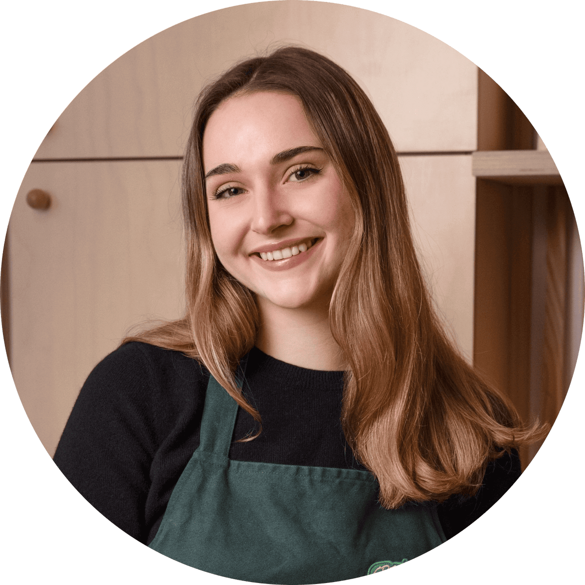 A young woman with long brown hair smiles at the camera, wearing a dark sweater and a green apron, standing in front of a light wooden background.