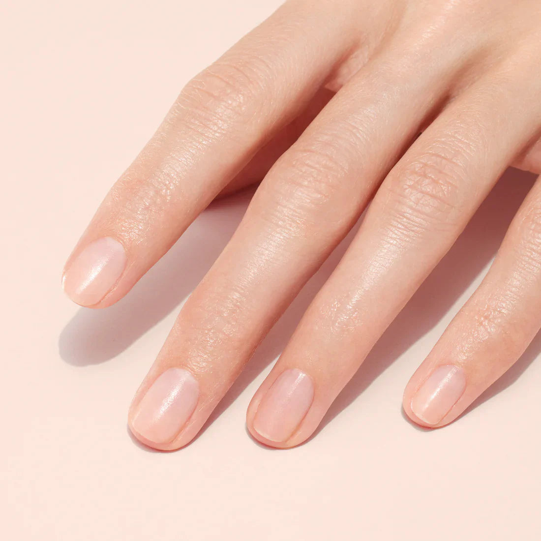 A close-up of a hand with neatly manicured, short natural nails on a light pink background. The nails are clean, shiny, and unpolished.