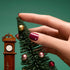 Une main à la manucure bordeaux impeccable, ornée des strass Nail Art Holidays de Manucurist, décore un petit sapin de Noël avec des ornements, pour une touche festive inspirée des looks de fêtes. Une horloge comtoise miniature se tient à côté du sapin, le tout sur fond vert.