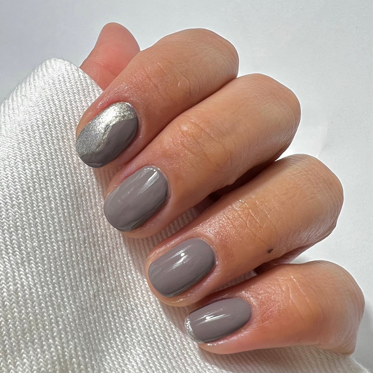 A hand with short, neatly manicured nails painted in glossy taupe polish. The index fingernail features a silver metallic accent design near the cuticle. The person is wearing a white knit sleeve.