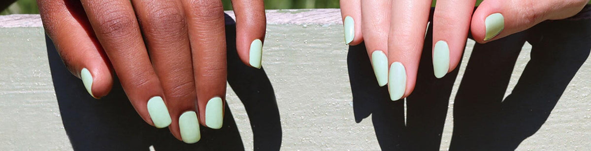 Two hands with light green matte nail polish rest on a light green wooden surface; one hand has a darker skin tone, and the other has a lighter skin tone.