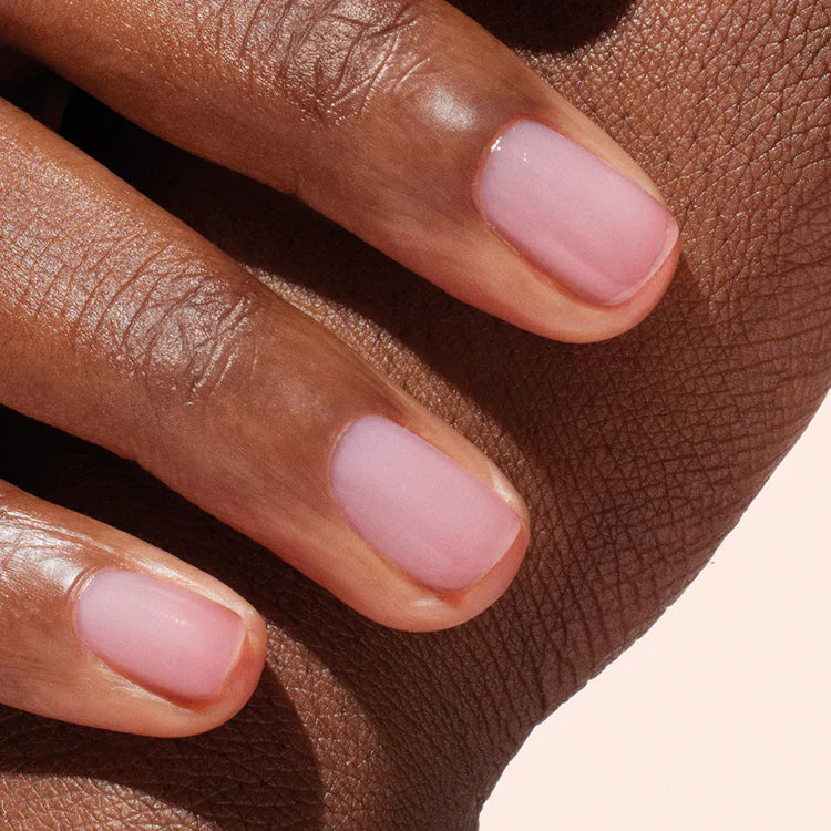 A close-up of a hand with medium-brown skin tone, showing neatly manicured nails painted with a sheer light pink polish. The hand rests gently on textured skin in the background.
