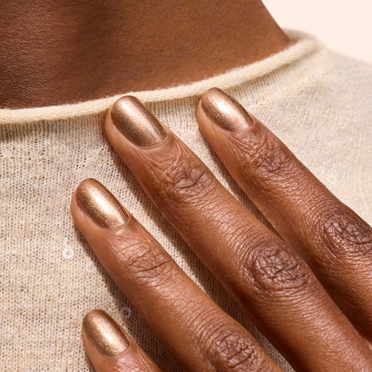 A close-up of a hand with Manucurist Bronzé gel polish on smooth, well-manicured nails, resting on a beige knit sweater and highlighting rich brown skin.
