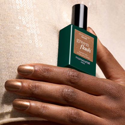 A hand with metallic bronze nail polish holds a green bottle of Manucurist Bronzé gel polish against a beige, sequined fabric background.