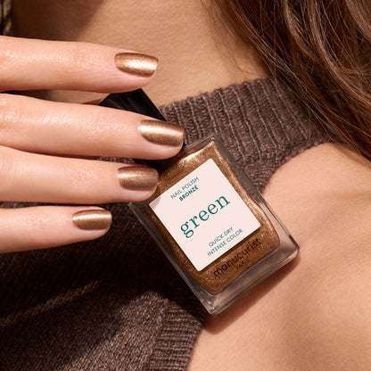 A hand with Bronzé, a bronze metallic nail polish by Manucurist, holds a matching bottle labeled "Manucurist Bronzé" against a brown knit sweater background.