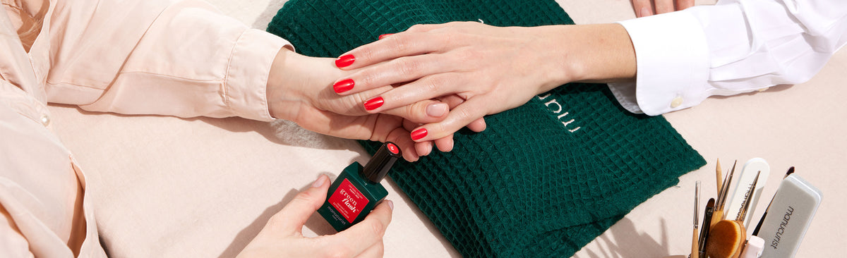 A person applies bright red nail polish to another persons fingernails, who is resting their hand on a green textured towel. Various manicure tools are visible on the side.