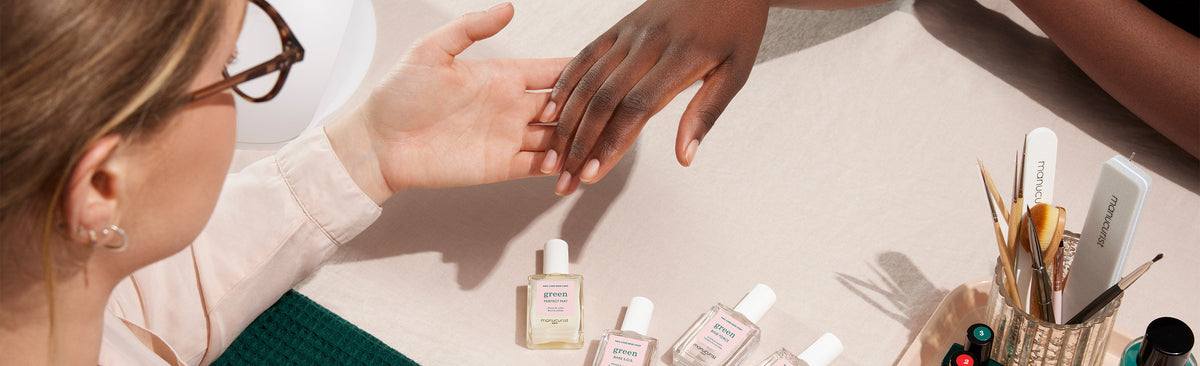 A manicurist wearing glasses holds a client’s hand while preparing to apply nail polish. Bottles of polish, manicure tools, and brushes are arranged on a beige table.