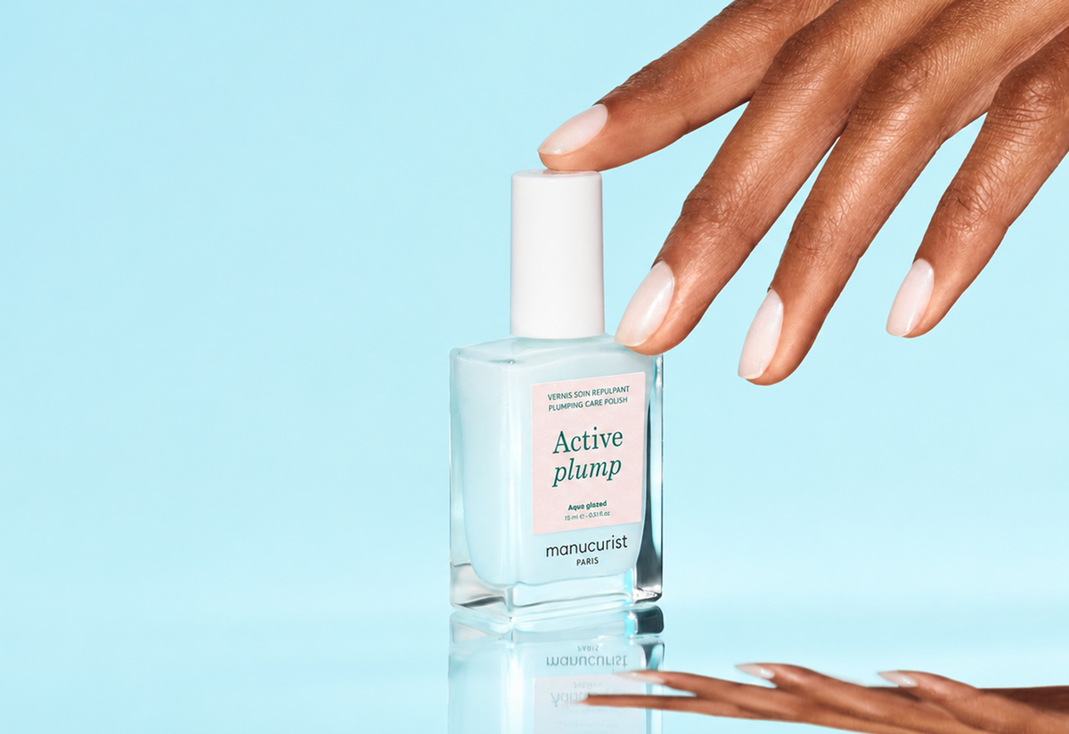 A hand with manicured nails reaches for a clear bottle of nail polish labeled Active Plump by Manucurist Paris, set against a light blue background.