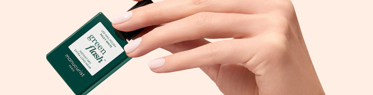 A hand with light pink nails holds a green bottle of Green Flash nail polish by Manucurist against a beige background.