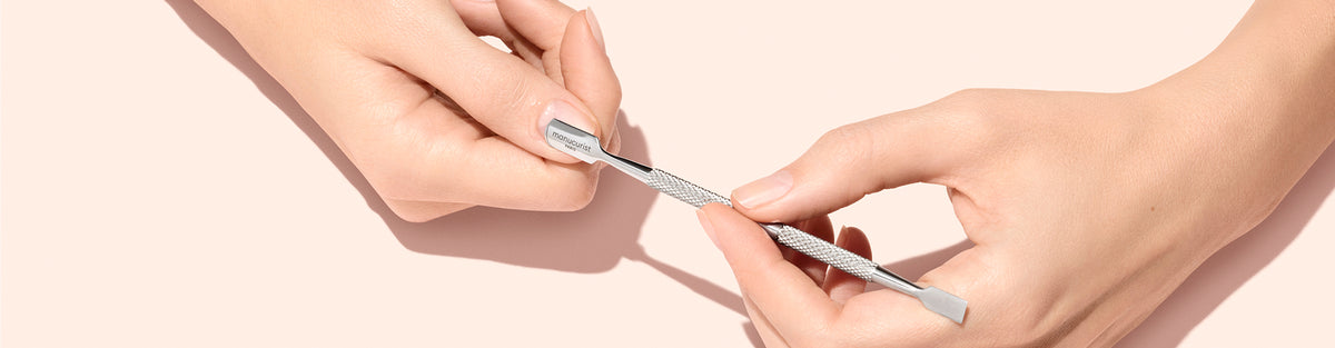 Two hands hold a metal cuticle pusher against a light beige background, with one hand grasping each end of the tool.