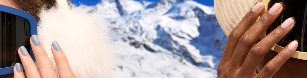Two people wearing winter earmuffs and sunglasses stand in front of a snowy mountain landscape. One has pale blue nails, the other has nude nails, both holding their sunglasses with hands near their faces.