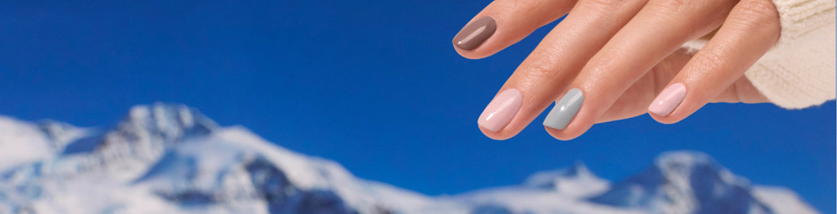 A hand with pastel-colored manicured nails is held up against a clear blue sky and snowy mountain peaks in the background. The person wears a cream-colored sweater.