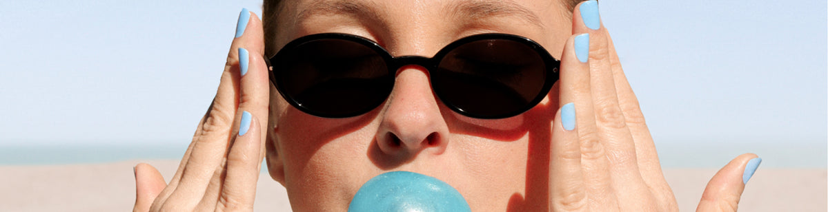 A person wearing black oval sunglasses and light blue nail polish holds their hands to their face while blowing a blue bubblegum bubble, with a blurred beach background.