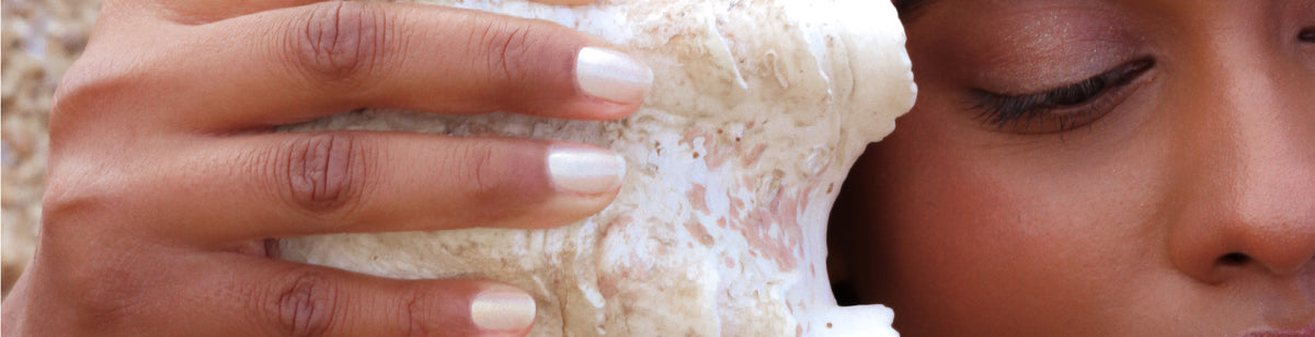 A close-up of a persons face and hand holding a large seashell to their ear, with the persons eye closed and nails painted a pale color.