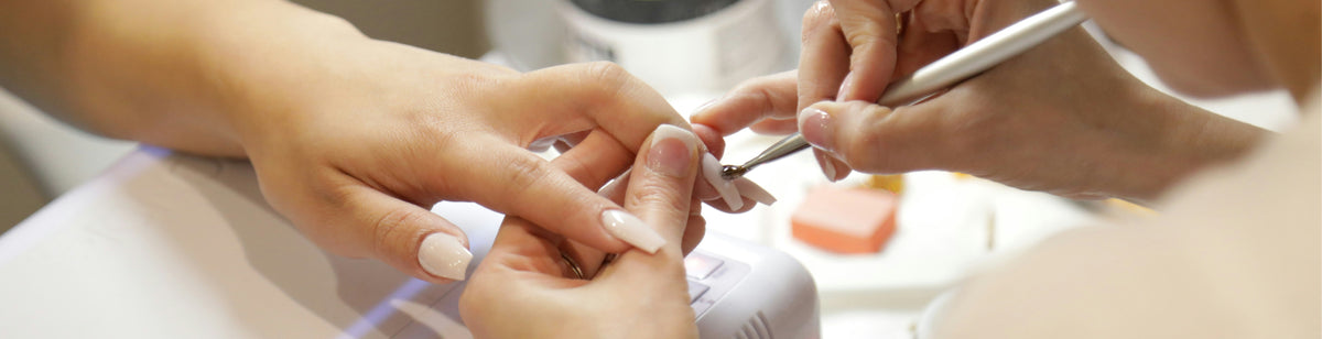 A close-up of a nail technician carefully painting a clients fingernails with a fine brush. The clients hand rests on a surface, and both hands have neatly manicured, light-colored nails.