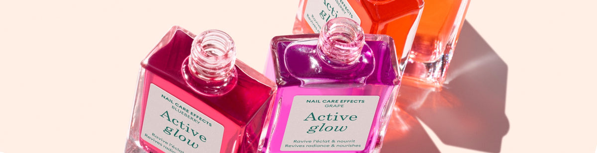 Three colorful glass bottles of nail care products labeled Active glow are tipped over, with one pink, one purple, and one orange bottle casting shadows on a light pink surface.