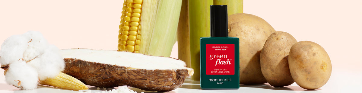 A bottle of Green Flash nail polish is placed among natural ingredients: cotton, a sliced cassava root, corn, baby corn, and potatoes, all arranged on a light surface with a soft pink background.