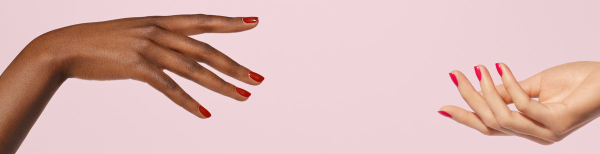 Two hands with manicured nails, one with dark skin and red polish, the other with light skin and pink polish, reaching toward each other against a pale pink background.