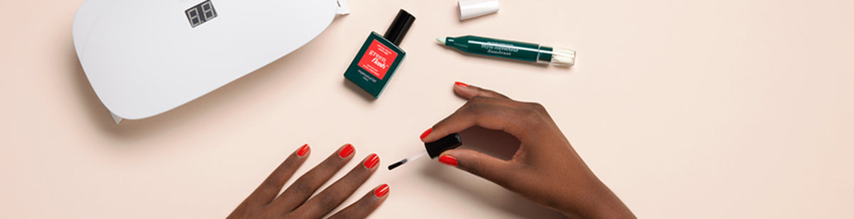 A person with dark skin applies clear nail polish to their red-painted nails at a table. Nearby are a white LED nail lamp, a bottle of nail polish, and a green correction pen with its cap removed.
