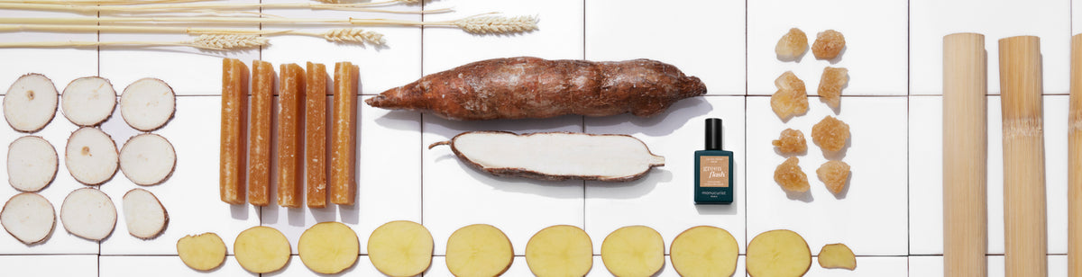 Various natural ingredients, sliced roots, sugarcane, golden chunks, wheat stalks, and a small dark green bottle are arranged neatly on a white tiled surface.