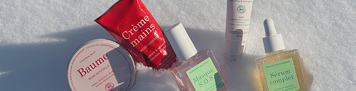 Five skincare products, including creams, serums, and a red hand cream tube, are arranged upright on white snow, with sunlight highlighting their packaging.