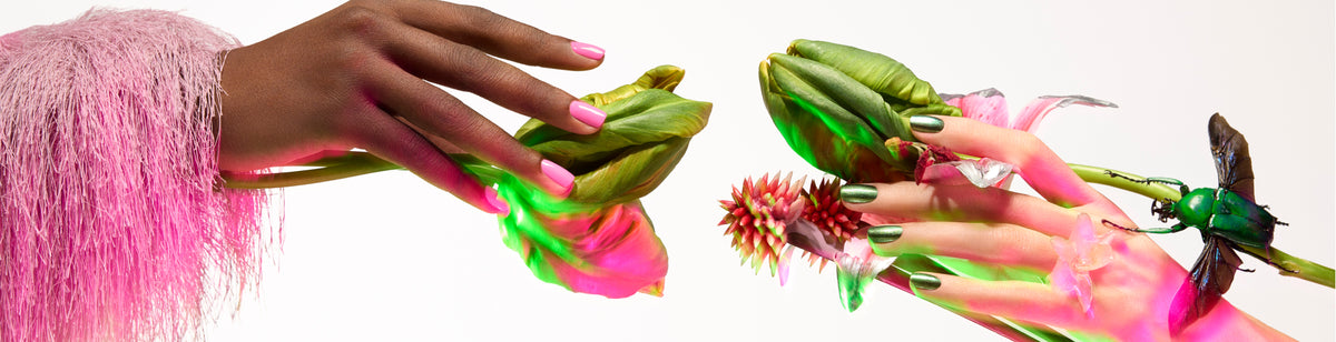 Two hands with painted nails touch green leaves and flowers. One hand is brown with pink nails and a fuzzy pink sleeve; the other is light-skinned with green nails and a beetle, both lit by pink and green light.