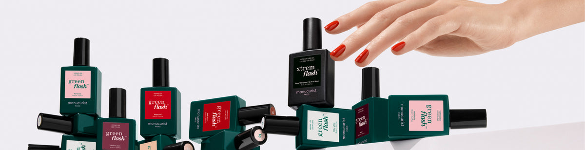 A manicured hand with red nail polish picks up a bottle of green flash nail polish among several scattered bottles of different colors and labels on a white surface.