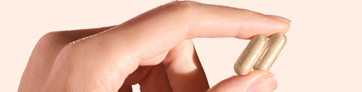 A close-up of a hand gently holding two beige capsules between the thumb and index finger against a light background.