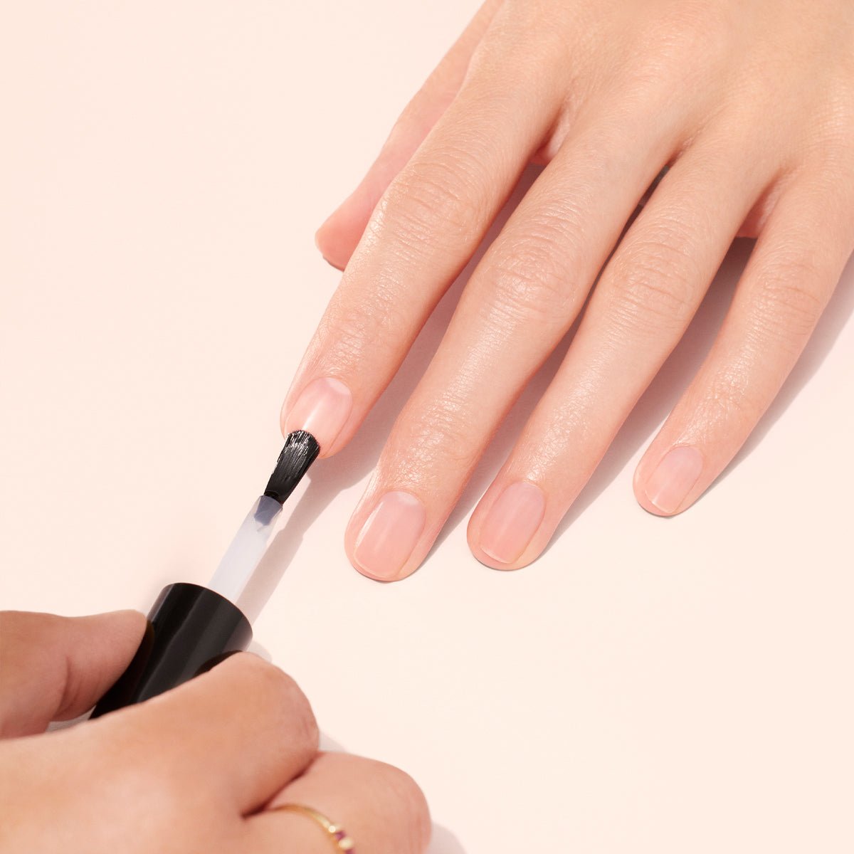 A person applies clear nail polish to the fingernails of another hand resting on a light pink surface.