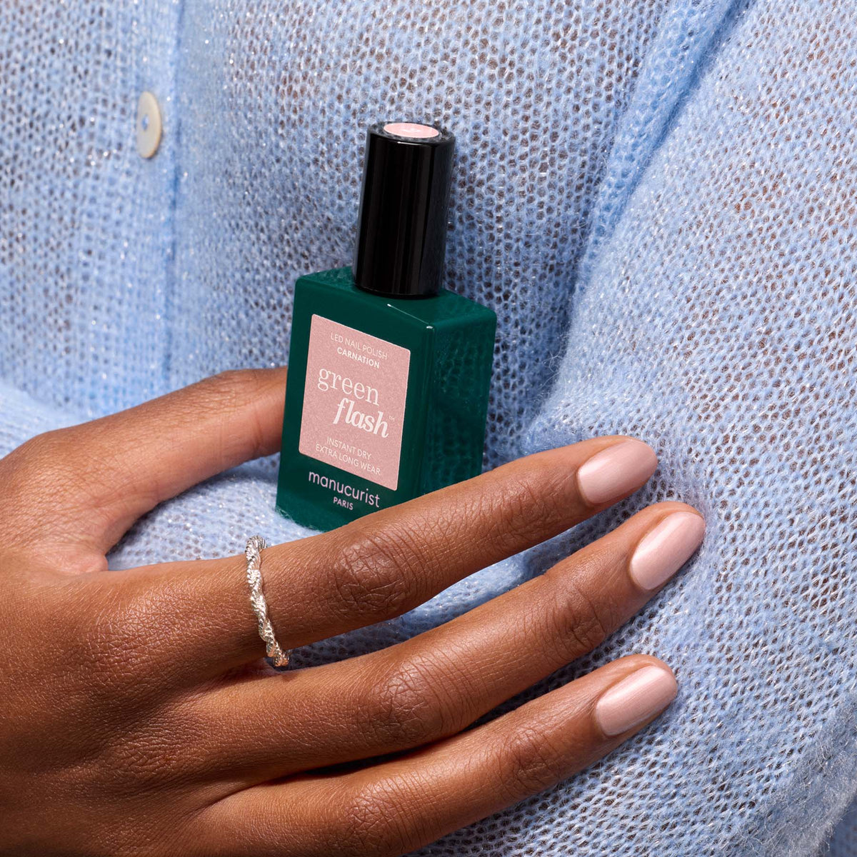 A person in a light blue knit sweater holds a bottle of Manucurist Carnation gel polish. Their nails are manicured in a pearly pink nude shade, accented by a delicate silver ring on their finger.