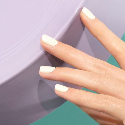 Une main aux ongles soigneusement manucurés et peints avec Porcelaine de Manucurist, une teinte blanc cassé crémeuse, repose sur une surface lisse violet clair devant un fond bleu sarcelle.