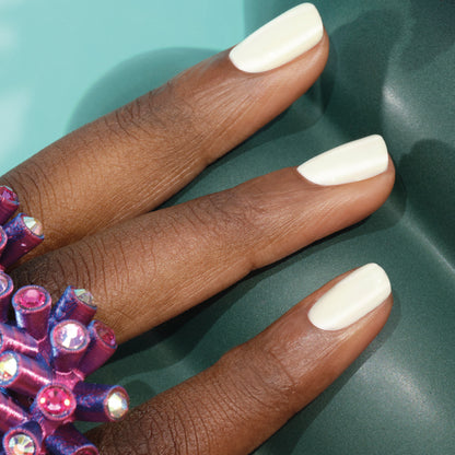 Gros plan sur une main à la peau foncée et lisse, aux ongles courts peints avec le vernis à ongles Porcelaine de Manucurist. La main porte une grosse bague violette ornée de gemmes rondes brillantes, sur un fond vert sarcelle.